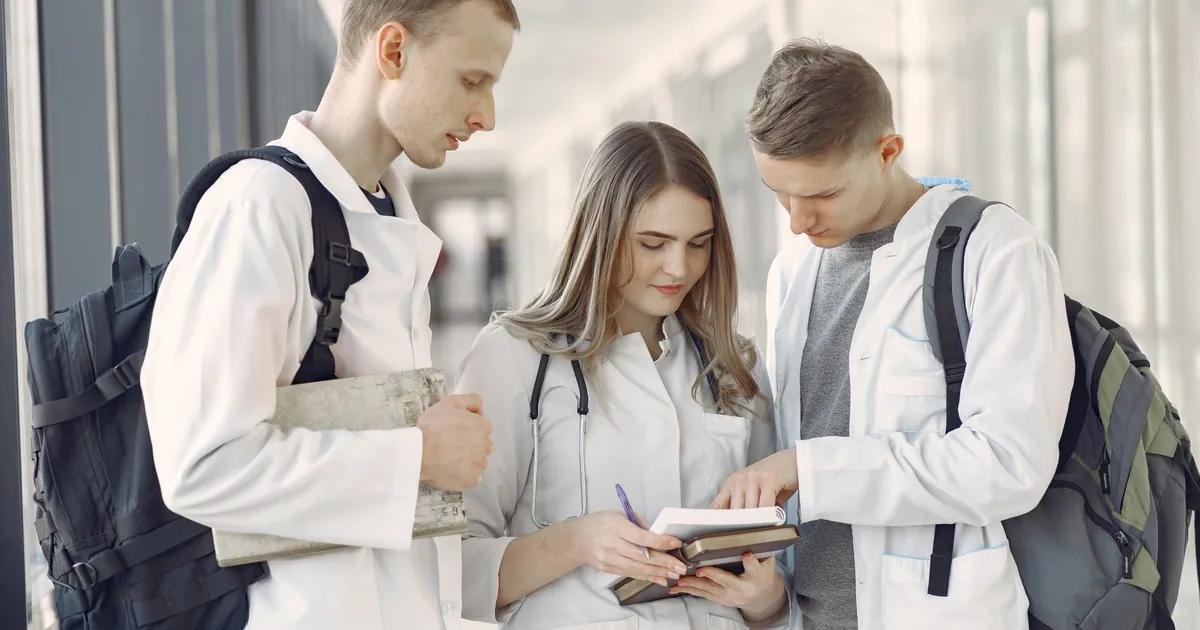 Three medical students in white coats reading notes together in a hospital corridor